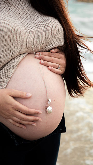 Une personne enceinte tient son ventre, portant un pendentif. Ses mains reposent tendrement dessus, près de la mer en arrière-plan flou.